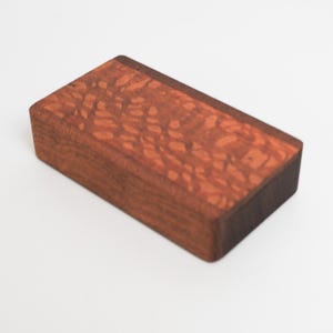 May include: A small wooden box with a reddish-brown finish and a dark brown edge. The wood has a unique grain pattern.