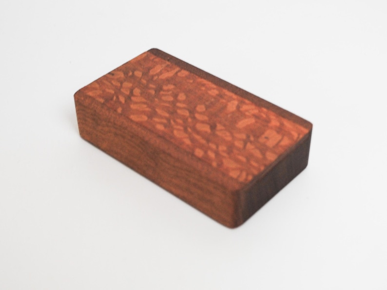 May include: A small wooden box with a reddish-brown finish and a dark brown edge. The wood has a unique grain pattern.