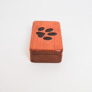 May include: A small wooden box with a paw print design. The box is made of cherry wood and has a natural finish.