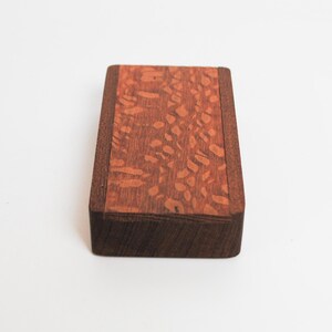 May include: A small wooden box with a dark brown border and a light brown top with a patterned design.