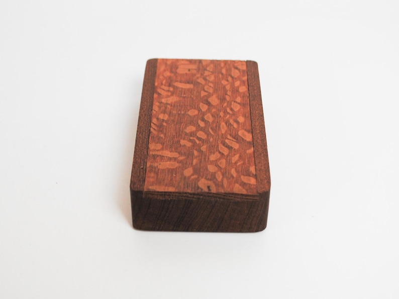 May include: A small wooden box with a dark brown border and a light brown top with a patterned design.