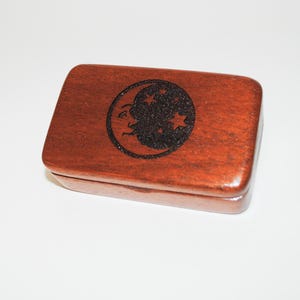 May include: A small wooden box with a crescent moon and stars design engraved on the lid. The box is made of dark brown wood and has a hinged lid.