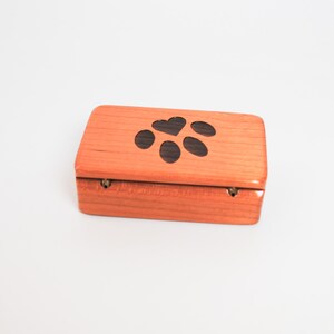 May include: A small wooden box with a hinged lid. The top of the box is decorated with a black paw print with a heart in the centre.