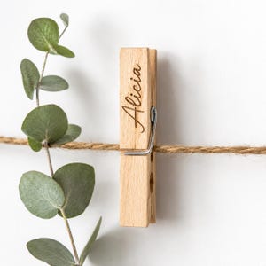 Könnte beinhalten: Eine Holz-Wäscheklammer mit dem Namen "Alicia" in kursiver Schrift graviert. Die Klammer ist an einer natürlichen Schnur befestigt, mit grünen Eukalyptusblättern im Hintergrund. Die Wäscheklammer ist aus hellem Holz.