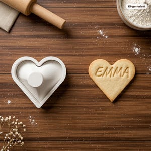 May include: A white heart-shaped cookie cutter with a central handle and a matching heart-shaped cookie stamped with the name EMMA. The items are arranged on a dark wood surface alongside a wooden rolling pin and a small bowl of white flour.
