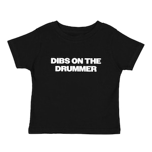 以下が含まれることがあります： 前面に白い文字「DIBS ON THE DRUMMER」が入った黒い幼児用Tシャツ。 このシャツは半袖のクルーネックで、カジュアルウェアに適しています。 テキストは太字のサンセリフ体です。