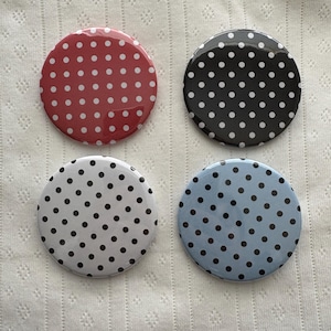 May include: Four round buttons with a polka dot pattern. The buttons are red with white dots, black with white dots, white with black dots, and light blue with black dots. The buttons are arranged on a white, textured surface.
