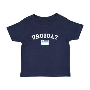 May include: Navy blue t-shirt featuring the word "URUGUAY" in white arched lettering above a small Uruguayan flag. The shirt is a short-sleeved crew neck style, suitable for children.