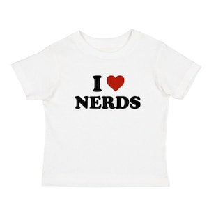 Puede incluir: Camiseta blanca con el texto "I ♥ NERDS" en letras negras y un corazón rojo. La camiseta es de manga corta y parece estar hecha de algodón. El diseño está centrado en la parte delantera de la camiseta.