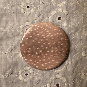 May include: A round, brown button with a white spotted pattern, resembling deer fur. The button is set against a light gray fabric with a floral embroidered design.