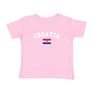 May include: A light pink t-shirt with the word "CROATIA" in white arched letters above the Croatian flag. The flag is a red, white, and blue checkered design with a coat of arms.