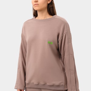 May include: A taupe-colored sweatshirt and matching pants set. The long-sleeved sweatshirt features a crew neck, decorative stitching on the sleeves, and a small green graphic on the chest. The pants have a matching green graphic.