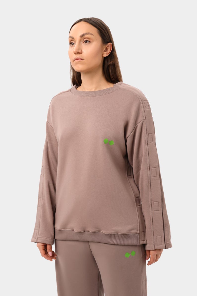 May include: A taupe-colored sweatshirt and matching pants set. The long-sleeved sweatshirt features a crew neck, decorative stitching on the sleeves, and a small green graphic on the chest. The pants have a matching green graphic.
