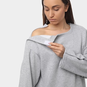 May include: A light gray sweatshirt with a unique design. The sweatshirt features a shoulder opening with a white velcro closure. The long sleeves have a relaxed fit. The model is wearing the sweatshirt.