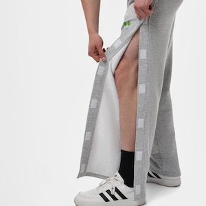 Op de afbeelding: Grijze joggingbroek met drukknoopsluitingen aan de zijkanten. De broek is open en toont een witte binnenkant. De persoon draagt witte sneakers met zwarte strepen en zwarte sokken. De broek heeft een klein groen logo bovenaan.