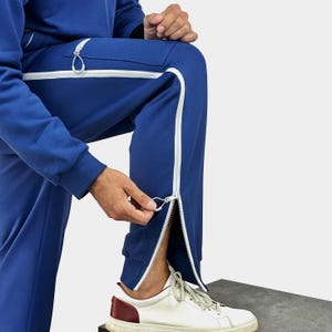 Op de afbeelding: Koning blauwe joggingbroek met witte ritsen aan de zijkanten. De broek heeft een bijpassend blauw trekkoord en een witte ritstrekker. De persoon draagt witte sneakers met bordeauxrode accenten.
