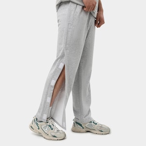 Op de afbeelding: Lichtgrijze joggingbroek met zijsplitten vastgemaakt met witte klittenbandsluitingen. De broek heeft een relaxte pasvorm en verticale naaddetails. De persoon draagt witte en groene sneakers.