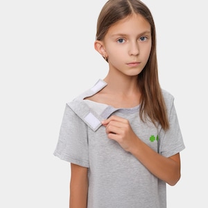 Puede incluir: Una camiseta gris de manga corta con un diseño único. La camiseta presenta un cuello que se abre con dos cierres de velcro blancos. Un pequeño diseño de cruz verde es visible en la parte delantera. La modelo lleva la camiseta.