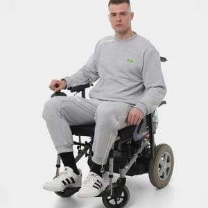 Puede incluir: Un conjunto de sudadera y pantalón de chándal gris claro. La sudadera tiene un pequeño diseño verde en el pecho y un detalle de cremallera en el hombro. La persona lleva zapatillas blancas con rayas negras y está sentada en una silla de ruedas.