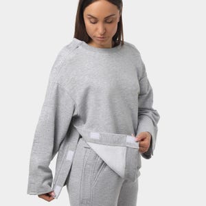 May include: A light gray sweatshirt and matching sweatpants set. The sweatshirt has a crew neck and wide sleeves. The front of the sweatshirt has a flap that is secured with velcro. The sweatpants have a drawstring waist and vertical seams.