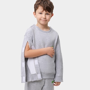 Puede incluir: Conjunto de sudadera y pantalón de chándal gris claro. La sudadera presenta un diseño único con mangas abiertas sujetas con cierres de velcro blancos. Un pequeño gráfico verde está en el pecho. Para niños.