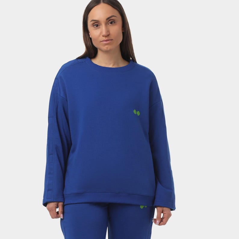 May include: A cobalt blue sweatshirt and matching pants set. The sweatshirt features small green clover-like designs on the chest and lower left side. The long sleeves have a subtle vertical stripe detail. The model is wearing the set.