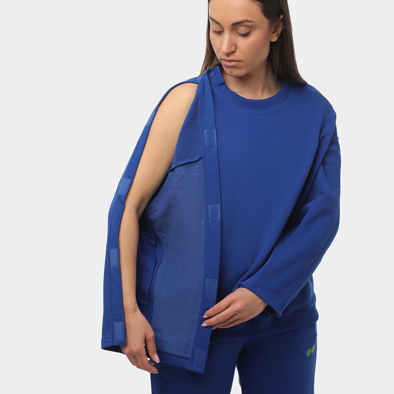 May include: A royal blue sweatshirt with a unique design. The sweatshirt features a split sleeve design with a series of velcro closures. The model is wearing matching blue pants. The overall style is modern and functional.