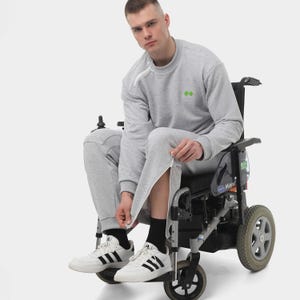 Puede incluir: Una persona en silla de ruedas con una sudadera gris y un pantalón de chándal a juego con cremallera. La sudadera tiene dos pequeños detalles bordados en verde. La persona lleva zapatillas blancas con rayas negras.