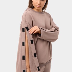 May include: A mauve-colored sweatshirt with long sleeves and a crew neck. The sleeves have a unique design with multiple black velcro closures. The model is wearing matching mauve sweatpants. The outfit is casual and comfortable.