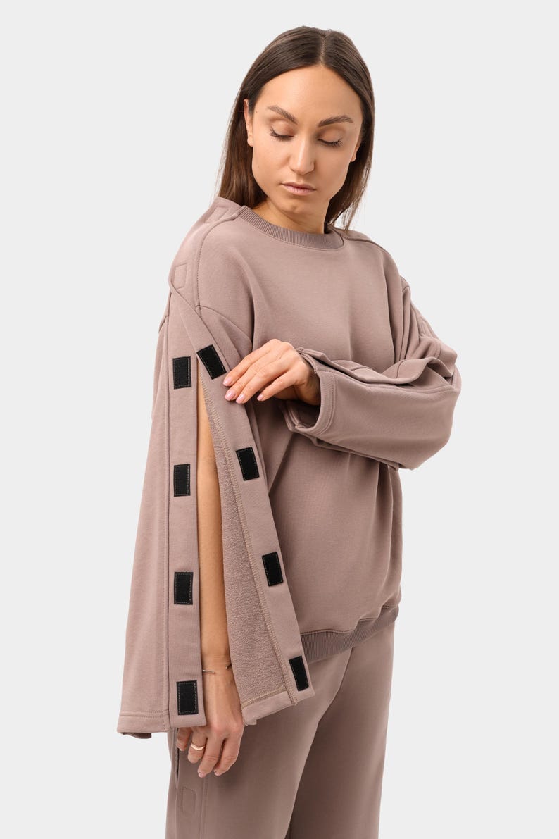 May include: A mauve-colored sweatshirt with long sleeves and a crew neck. The sleeves have a unique design with multiple black velcro closures. The model is wearing matching mauve sweatpants. The outfit is casual and comfortable.