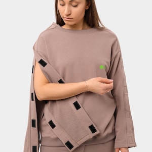 Puede incluir: Una sudadera color topo con mangas desmontables, aseguradas con cierres de velcro negros. Un pequeño trébol verde está en el pecho. Se ve un pantalón a juego. El conjunto es de estilo casual y moderno.