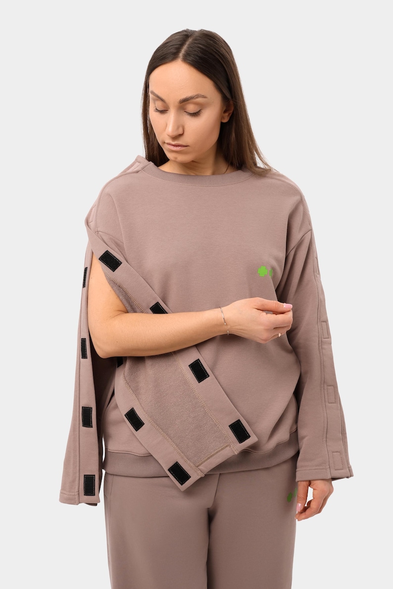 May include: A taupe-colored sweatshirt with detachable sleeves, featuring black velcro closures. A small green shamrock is on the chest. The matching pants are visible at the bottom. The outfit is a casual, modern style.