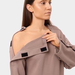 May include: A mauve-colored sweatshirt with a unique design. The garment features a wide, open neckline with a fold-over collar secured by black hook-and-loop fasteners. The sleeves have similar fasteners.