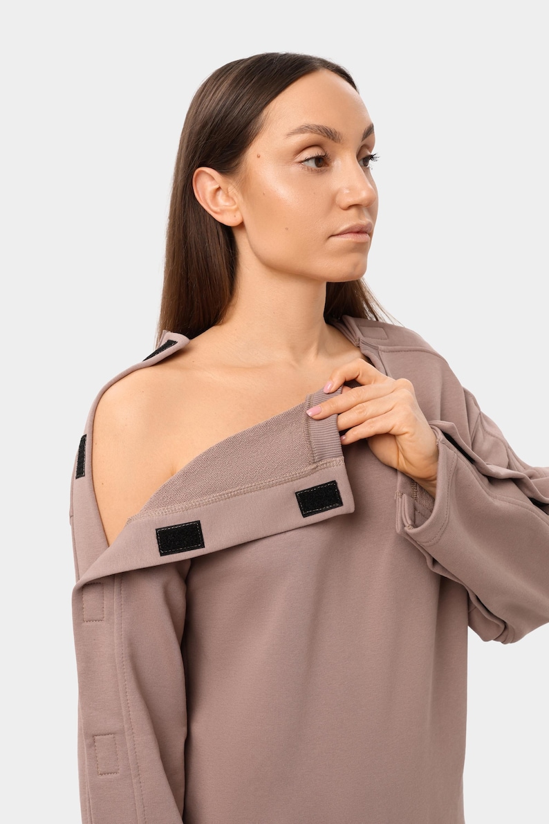 May include: A mauve-colored sweatshirt with a unique design. The garment features a wide, open neckline with a fold-over collar secured by black hook-and-loop fasteners. The sleeves have similar fasteners.