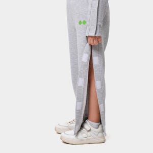 Puede incluir: Pantalones de chándal grises claros con una abertura lateral asegurada con parches de velcro blancos. Los pantalones tienen un pequeño diseño verde cerca de la cadera. Se ven zapatillas blancas con correas de velcro y calcetines blancos.