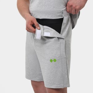 May include: Light gray shorts with a black waistband and white velcro closures. The shorts have a small green graphic on the left leg. The person in the image is adjusting the velcro closure.