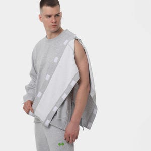 Op de afbeelding: Een grijze sweatshirt en bijpassende joggingbroek. De sweatshirt heeft een wit paneel over de borst en arm, vastgezet met witte klittenband vierkanten. De broek heeft een kleine groene afbeelding. Het model draagt de set.