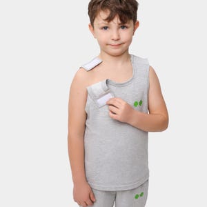 Puede incluir: Una camiseta sin mangas gris con una correa de velcro blanca en el hombro y el pecho. La camiseta tiene dos diseños de tréboles verdes en la parte delantera. El niño lleva pantalones cortos grises a juego con dos diseños de tréboles verdes.