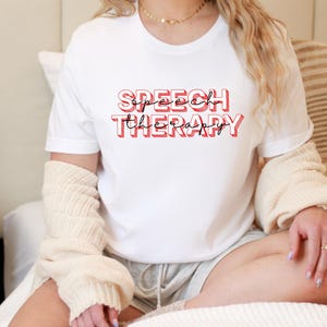 May include: White t-shirt with the words "SPEECH THERAPY" in red and black lettering. The shirt is short-sleeved and appears to be made of a soft material. The shirt is being worn.