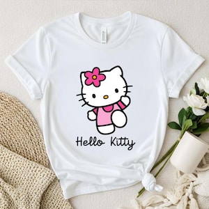 May include: White t-shirt featuring a Hello Kitty graphic. The cartoon cat is wearing a pink dress and a flower in her hair. The words "Hello Kitty" are written in a cursive font below the graphic.