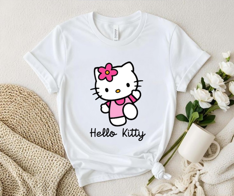 May include: White t-shirt featuring a Hello Kitty graphic. The cartoon cat is wearing a pink dress and a flower in her hair. The words "Hello Kitty" are written in a cursive font below the graphic.
