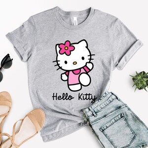 May include: A light gray t-shirt featuring a Hello Kitty graphic. The cartoon character is wearing a pink dress and a flower in her hair. The words "Hello Kitty" are printed in black below the graphic. The shirt is paired with denim shorts and sandals.