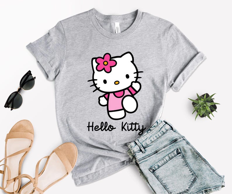 May include: A light gray t-shirt featuring a Hello Kitty graphic. The cartoon character is wearing a pink dress and a flower in her hair. The words "Hello Kitty" are printed in black below the graphic. The shirt is paired with denim shorts and sandals.
