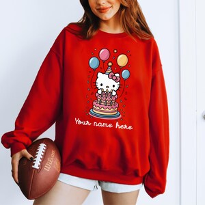 May include: Red sweatshirt featuring a cartoon character with a birthday cake and balloons. The graphic includes the text "Your name here". The sweatshirt is a long-sleeved, casual top.