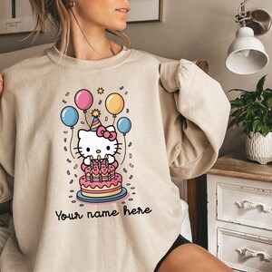 May include: Beige sweatshirt featuring a cartoon image of a white cat with a pink bow, a birthday cake, and balloons. The text "Your name here" is written in black script at the bottom of the design. The design is in a cartoon style.
