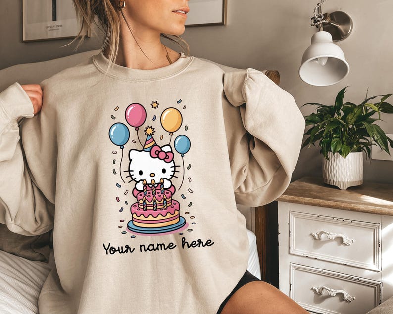 May include: Beige sweatshirt featuring a cartoon image of a white cat with a pink bow, a birthday cake, and balloons. The text "Your name here" is written in black script at the bottom of the design. The design is in a cartoon style.