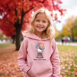 May include: A light pink hoodie featuring a graphic of a cartoon cat with a birthday cake, balloons, and the text "Your name here". The hoodie has a front pocket and a hood. The background shows a girl smiling in an outdoor setting.