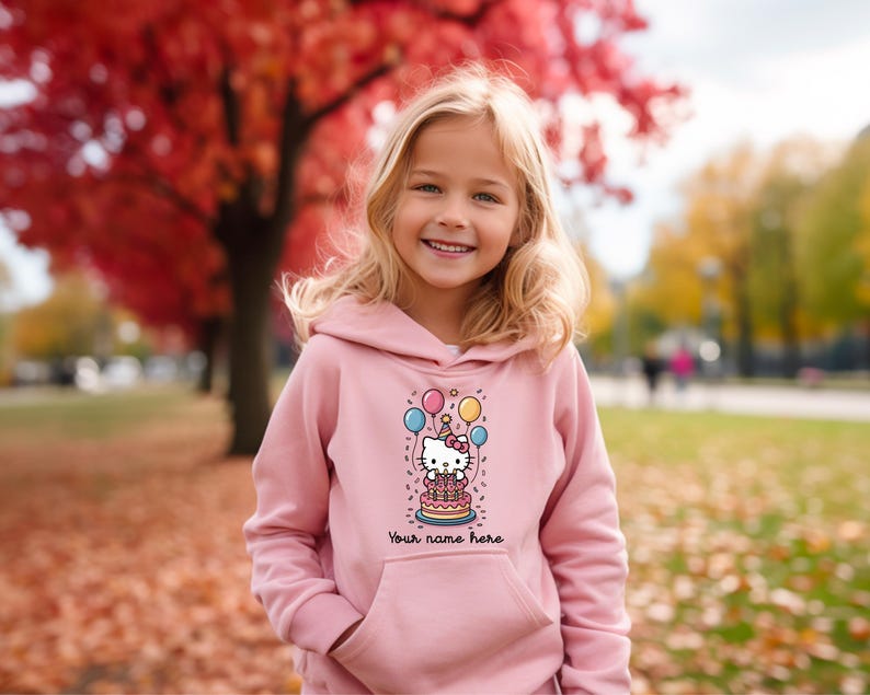 May include: A light pink hoodie featuring a graphic of a cartoon cat with a birthday cake, balloons, and the text "Your name here". The hoodie has a front pocket and a hood. The background shows a girl smiling in an outdoor setting.