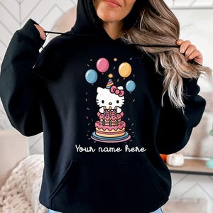 May include: Black hooded sweatshirt featuring a colorful graphic of a cartoon cat with a birthday cake, balloons, and confetti. The text "Your name here" is below the graphic. The sweatshirt has a front pocket and a drawstring hood.