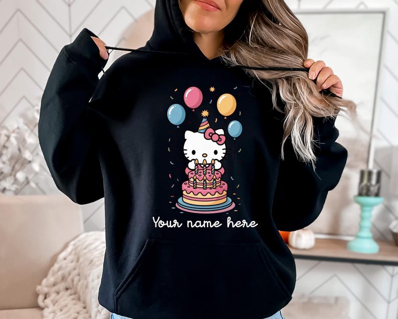 May include: Black hooded sweatshirt featuring a colorful graphic of a cartoon cat with a birthday cake, balloons, and confetti. The text "Your name here" is below the graphic. The sweatshirt has a front pocket and a drawstring hood.